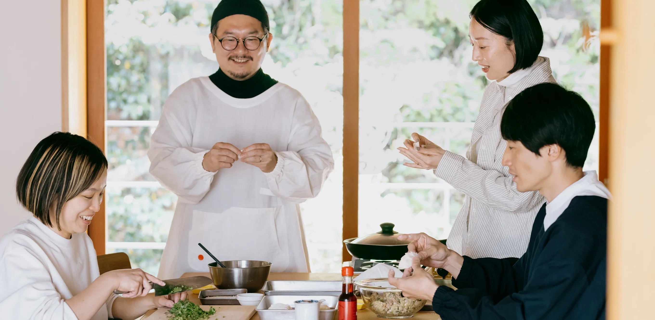 男女四人が仲良く餃子の皮を包んでいる様子。ホワイト色とネイビー色のみんなにいい割烹着を男性二人が着ている